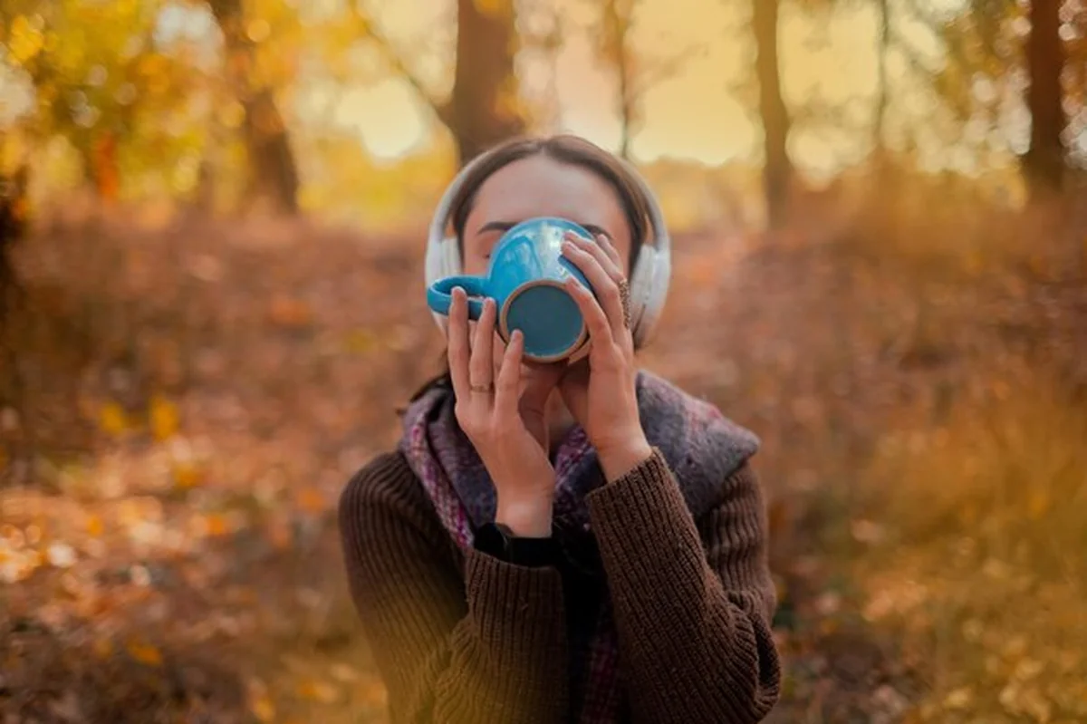 Kobieta z plecakiem pije herbatę, ubrana w brązowy sweter, trzyma mapę w rękach